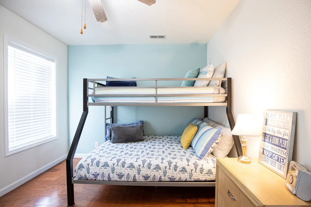 A playful and inviting secondary bedroom with a bunk bed, a cozy daybed, decorative pillows, and a nightstand with a lamp. The space features cheerful wall decor and creates an atmosphere suited for families or groups, highlighting flexible sleeping arrangements and a warm, welcoming mood.