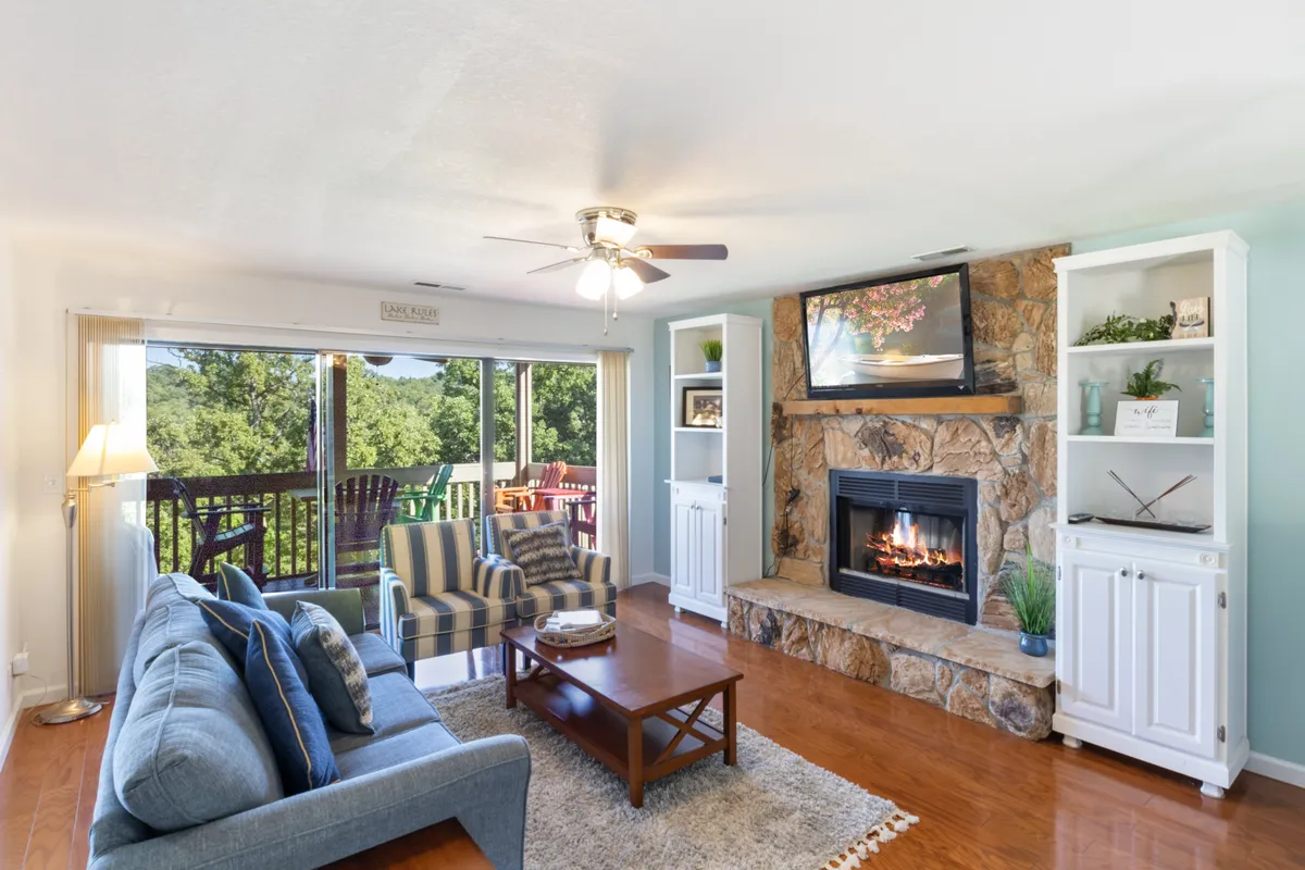 A cozy living room with a fireplace, comfortable sofa, bookshelves, and expansive windows overlooking the outdoors. The space invites relaxation, socializing, or enjoying the changing lake scenery through the seasons.