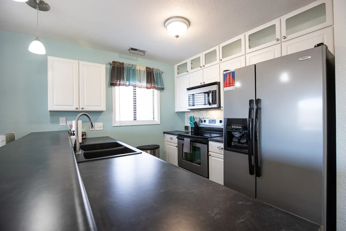 A modern kitchen scene with a prominent island, stainless steel refrigerator, oven, stove, and a window with curtains. The setup is open and functional, ideal for both cooking and entertaining guests, with a focus on practicality and togetherness.