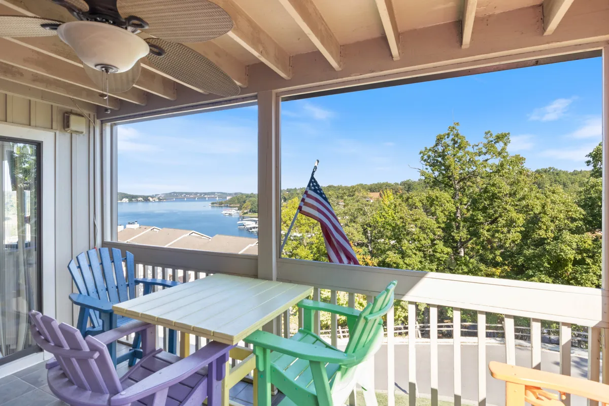 The image features a cheerful patio setup with colorful Adirondack chairs and an outdoor dining table overlooking a sparkling lake. An American flag waves gently in the breeze, and the view showcases tranquil water, inviting guests to relax, dine al fresco, and soak up the scenic surroundings.