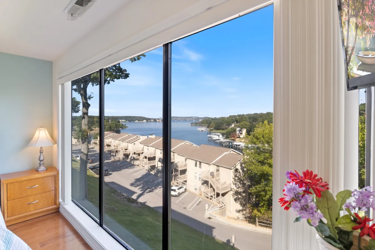 A bright interior shot with large windows framing a stunning lake view. The space features a decorative lamp, wooden dresser, and fresh flowers, creating a tranquil spot to relax and watch the sun set over the water.