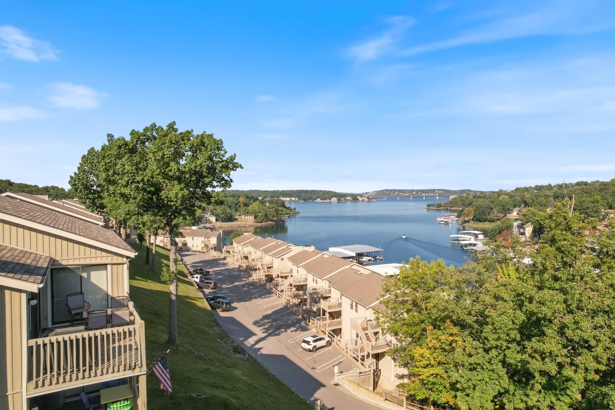 A panoramic lake view with boathouses along the shore, surrounded by lush trees and an expansive sky. The scene is tranquil, with soft reflections on the water and hints of lakeside activity such as walking paths and boating.