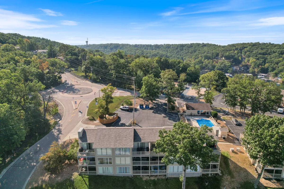 An exterior shot capturing the vibrant pool area surrounded by trees, with a building structure and roundabout visible in the background. The scene suggests opportunities for swimming, socializing, and enjoying nature within the community.