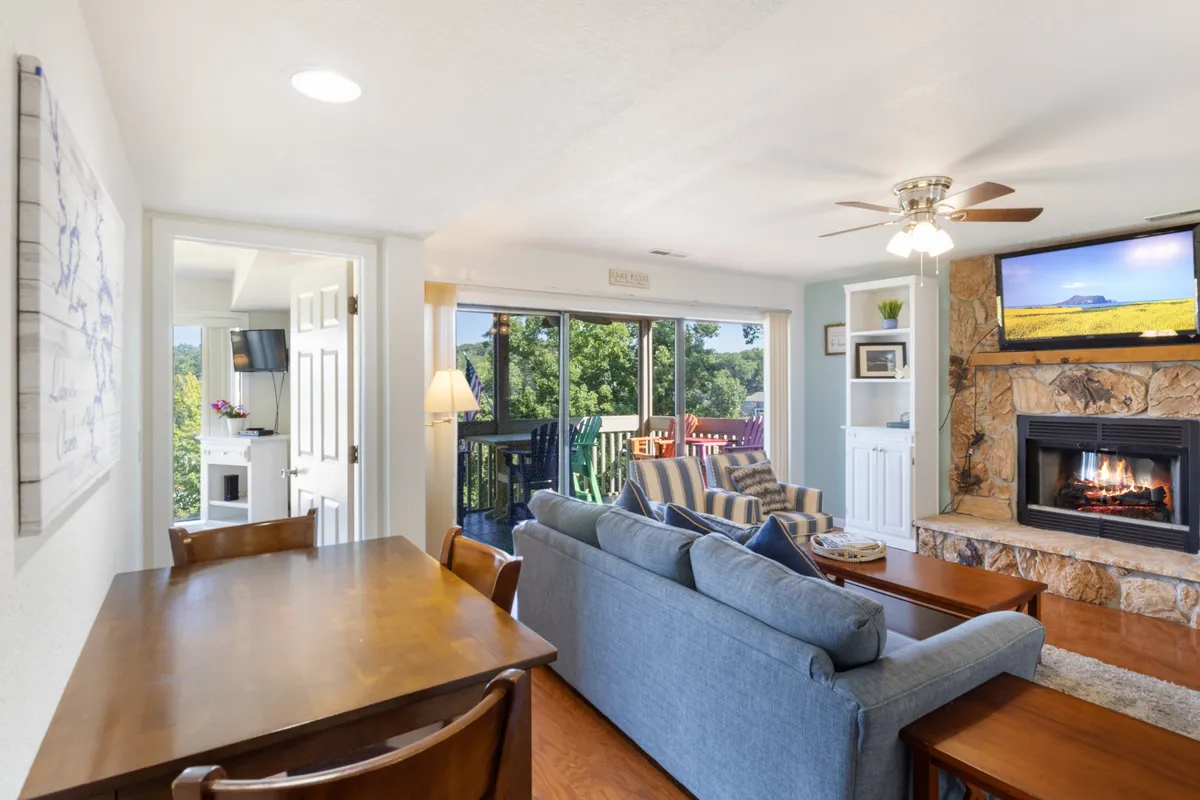 A welcoming open-concept living and dining area featuring a cozy fireplace, comfortable sofa, spacious dining table, and expansive windows that frame beautiful lake views. The arrangement invites gatherings, relaxation, and conversation, all bathed in natural light with direct sightlines to the outdoors.